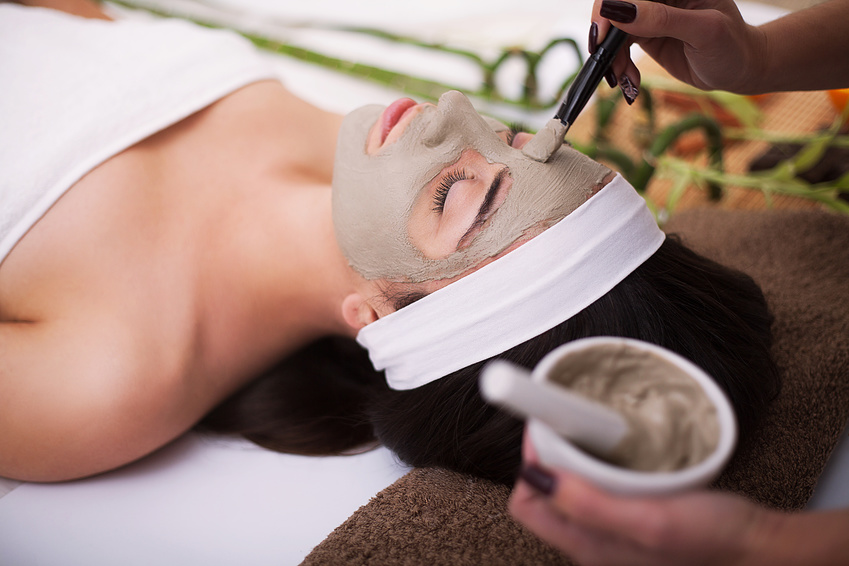 Spa. Beautiful young woman is getting facial clay mask at spa, lying with cucumbers on eyes